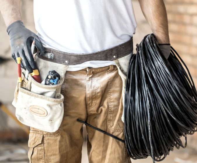 Ein Elektriker in Arbeitsklammotten hält ein langes Kabel in der Hand und in der anderen Werkzeuge. Er steht auf einer Baustelle und beschäftigt sich mit der Elektroinstallation.