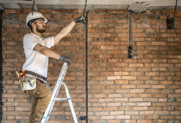 Elektriker aus Braunschweig steht auf einer Leiter und kürzt Kabel, die von der Decke hängen, um anschließend die Beleuchtung anzubringen.